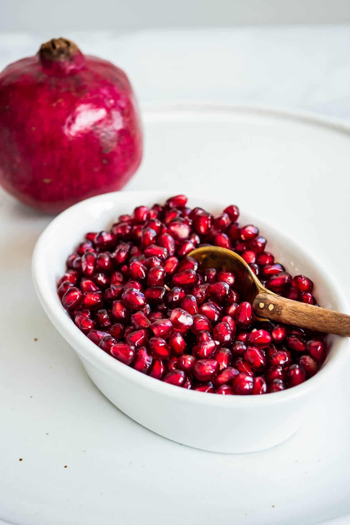 a close up of a bowl of pomegranate seeds in a bowl with a spoon, with a whole pomegranate in the background.