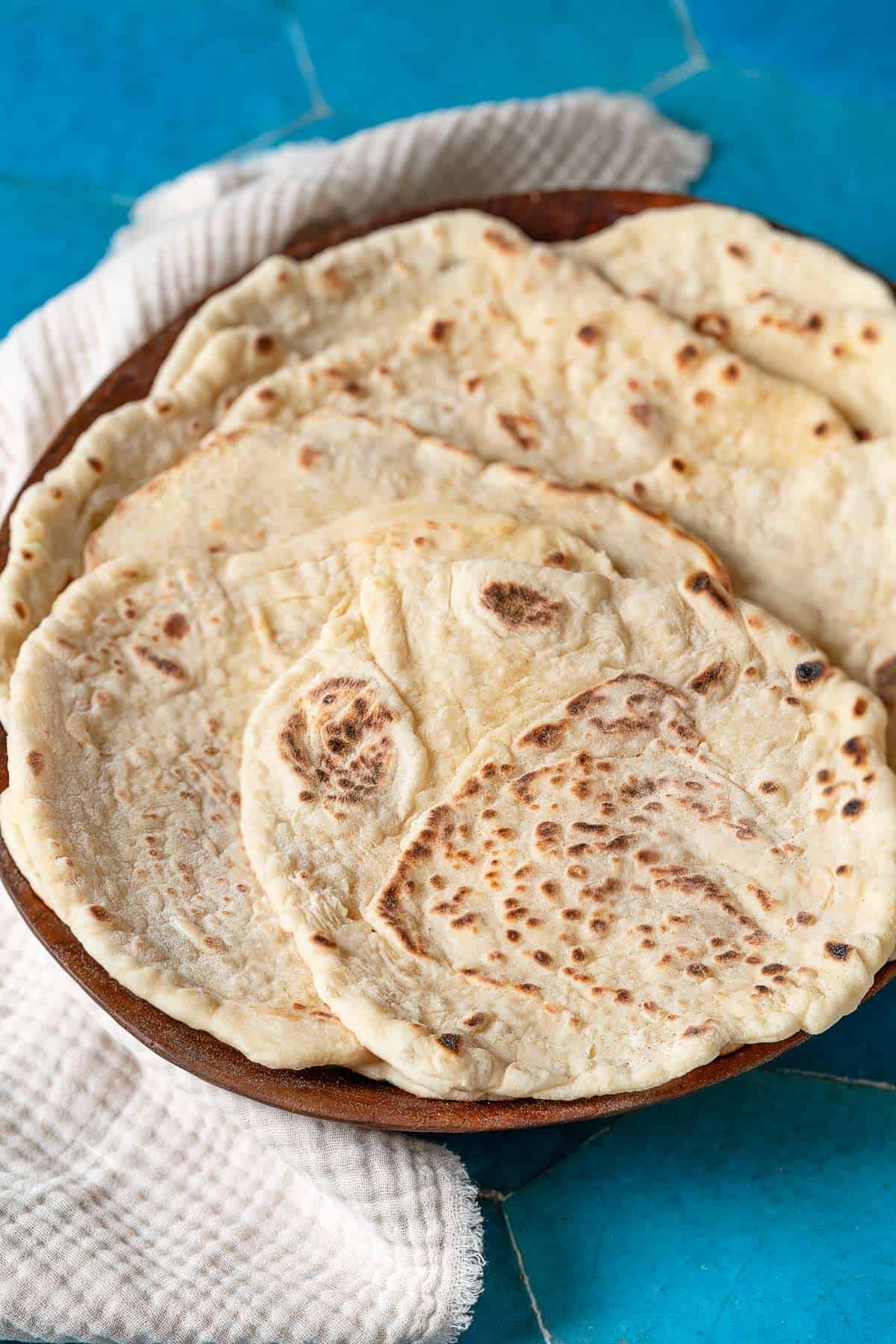 four lavash breads stacked in a brown bowl with a white linen cloth on the side.