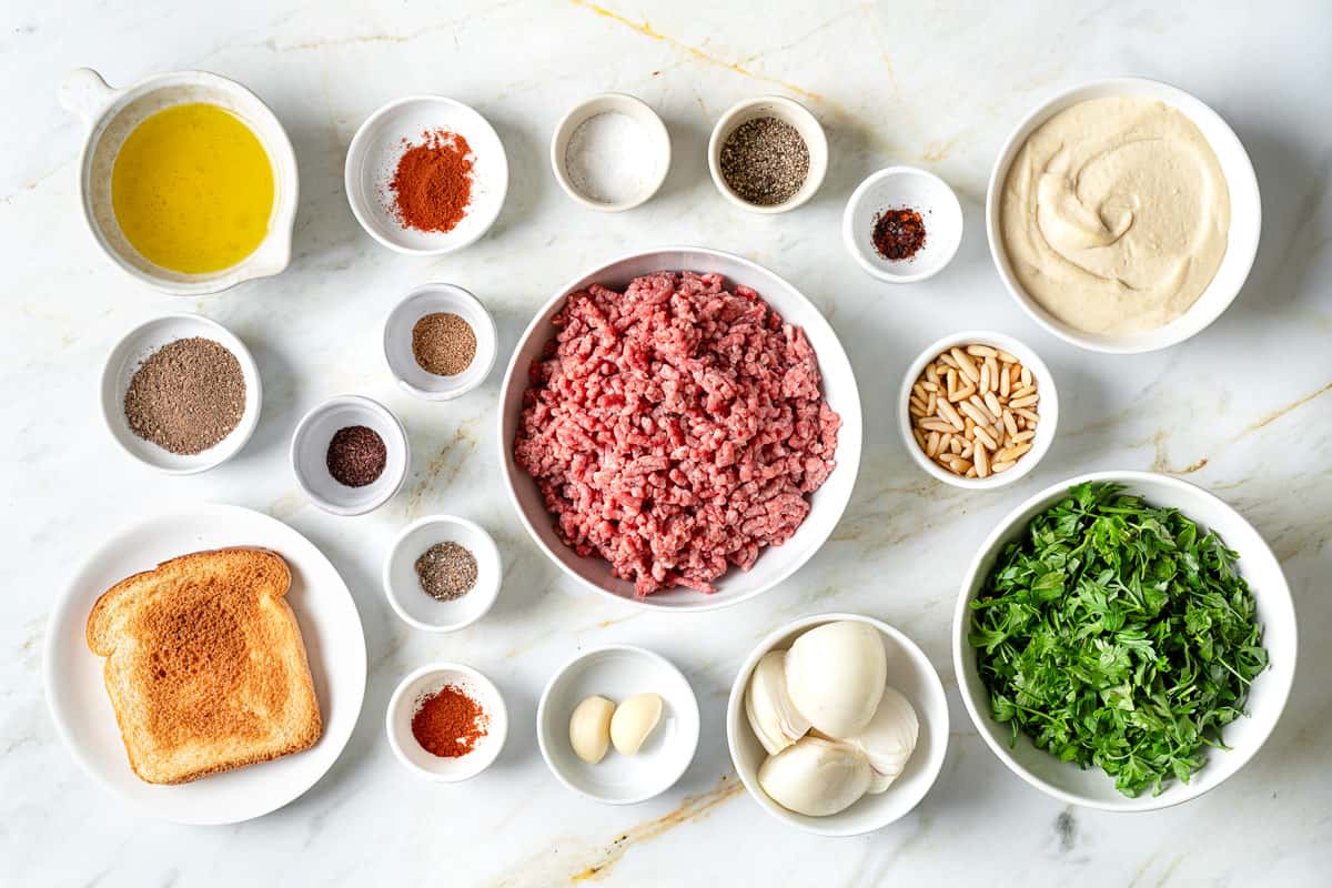 ingredients for meatballs over hummus including ground beef or lamb, bread, onion, garlic, parsley, salt, pepper, allspice, cardamom, sumac, nutmeg, paprika, cayenne, olive oil, pine nuts and hummus.