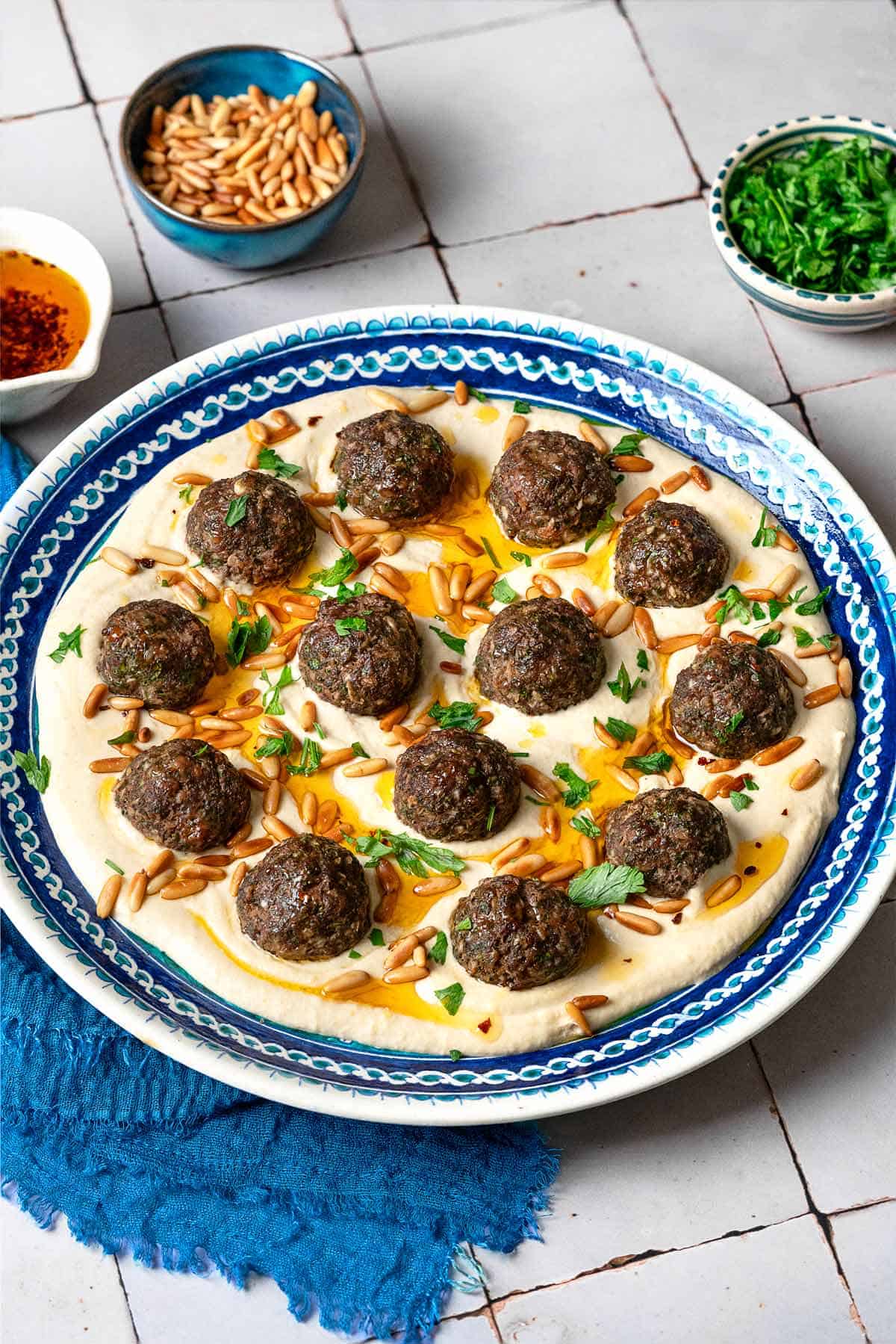 baked meatballs over hummus garnished with olive oil, pine nuts and parsley on a plate next to a blue napkin, and small bowls of parsley, pine nuts and olive oil.