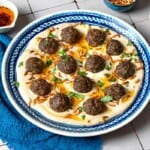 baked meatballs over hummus garnished with olive oil, pine nuts and parsley on a plate next to a blue napkin, a stack of blue plates with forks, and small bowls of parsley, pine nuts and olive oil.