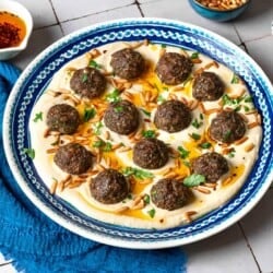 baked meatballs over hummus garnished with olive oil, pine nuts and parsley on a plate next to a blue napkin, a stack of blue plates with forks, and small bowls of parsley, pine nuts and olive oil.
