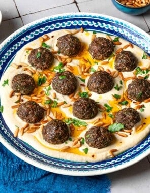 baked meatballs over hummus garnished with olive oil, pine nuts and parsley on a plate next to a blue napkin, a stack of blue plates with forks, and small bowls of parsley, pine nuts and olive oil.