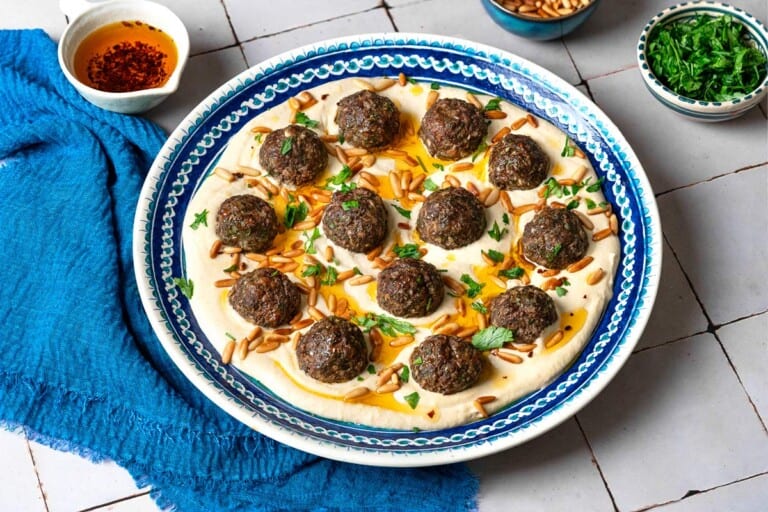 baked meatballs over hummus garnished with olive oil, pine nuts and parsley on a plate next to a blue napkin, a stack of blue plates with forks, and small bowls of parsley, pine nuts and olive oil.