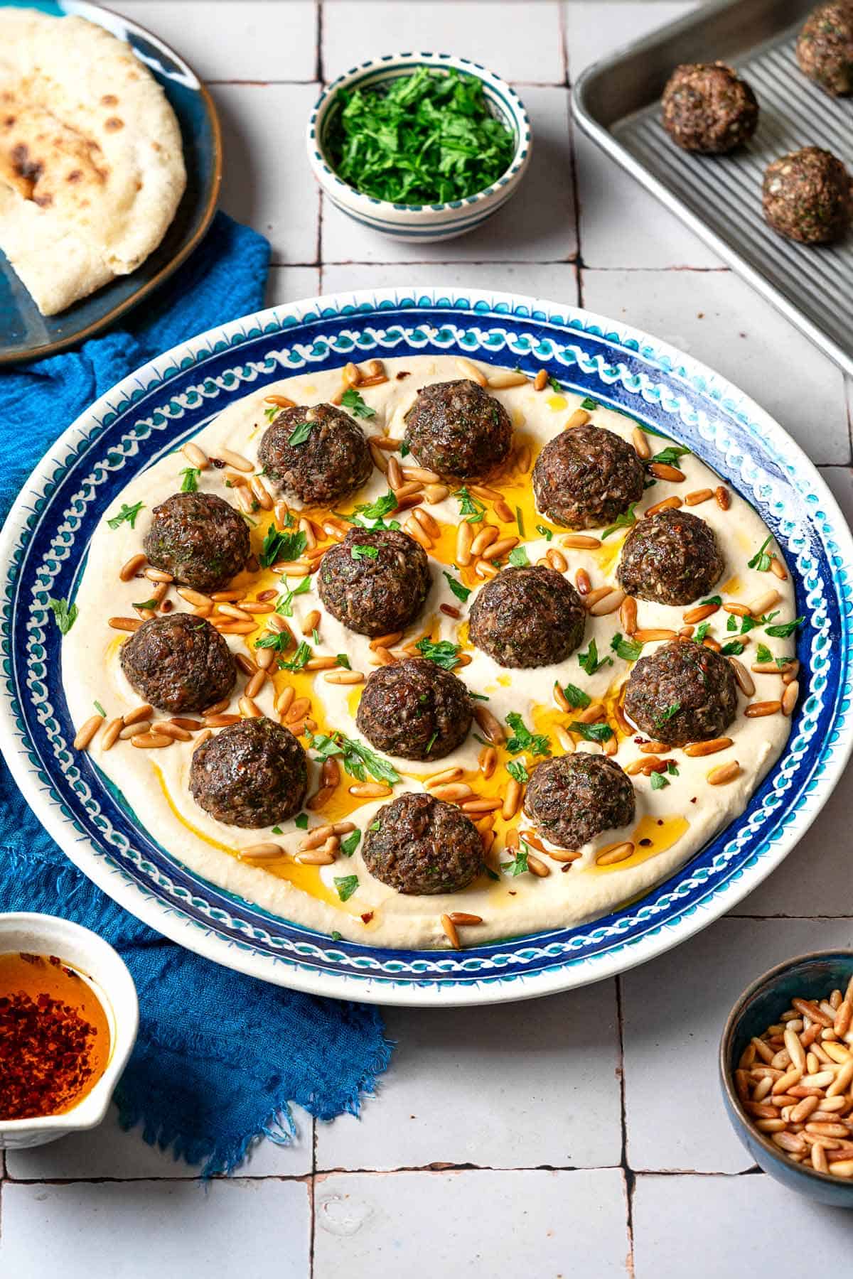 baked meatballs over hummus garnished with olive oil, pine nuts and parsley on a plate next to a blue napkin surrounded by a plate with pita, and small bowls of parsley, pine nuts and olive oil.