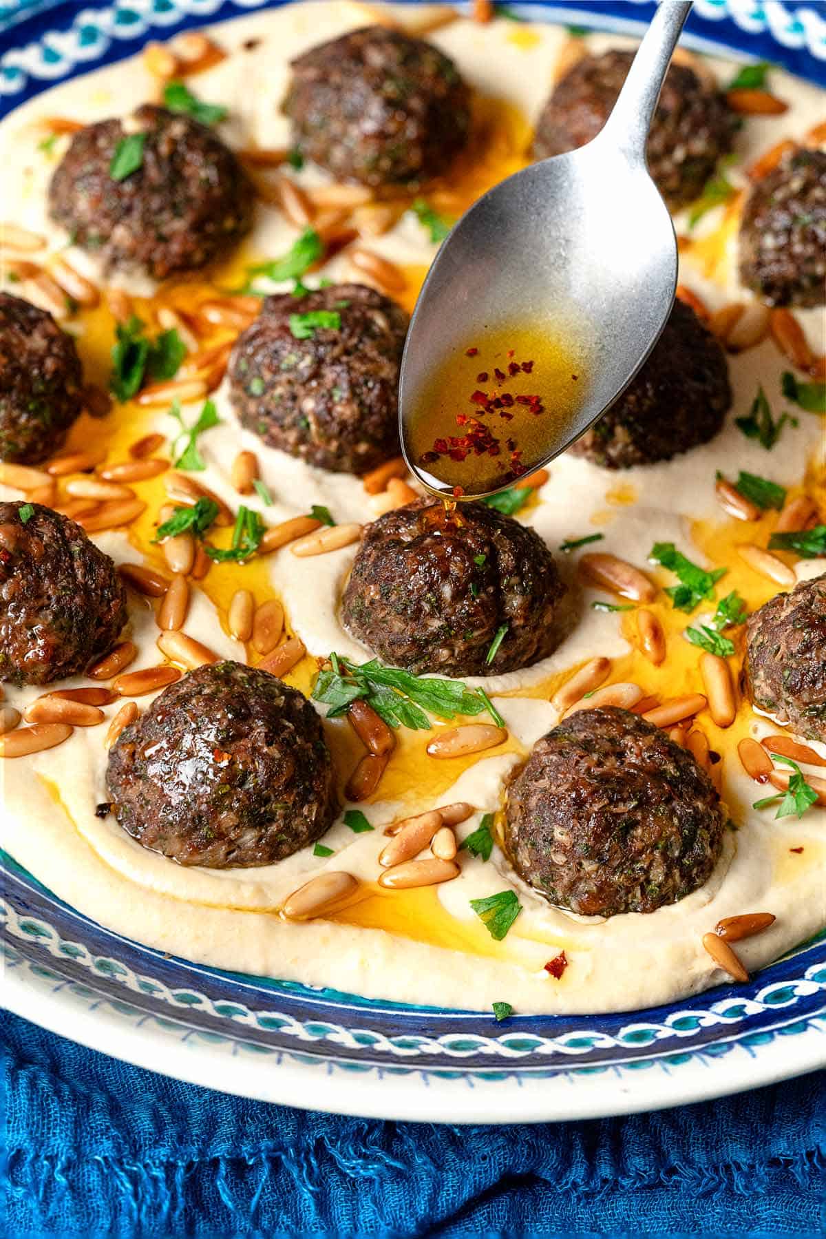 a close up of aleppo pepper infused olive oil being drizzled onto a plate of meatballs over hummus with a spoon.