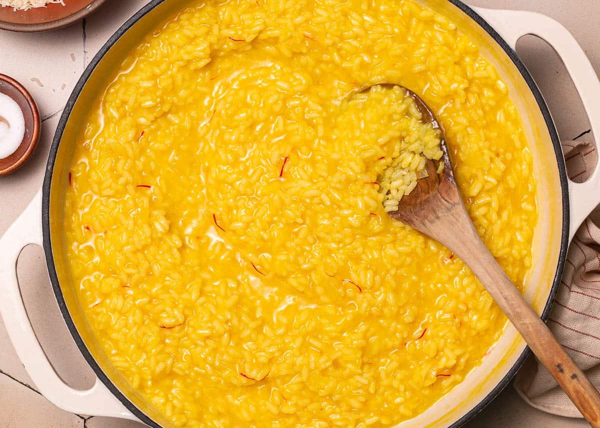 a close up of risotto alla milanese saffron risotto in a large pot being stirred with a wooden spoon.