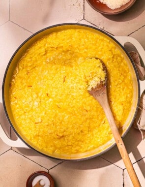 risotto alla milanese saffron risotto in a large pot being stirred with a wooden spoon next to a small bowl of salt with a spoon and a small bowl of grated parmesan cheese.