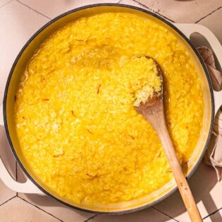 risotto alla milanese saffron risotto in a large pot being stirred with a wooden spoon next to a small bowl of salt with a spoon and a small bowl of grated parmesan cheese.