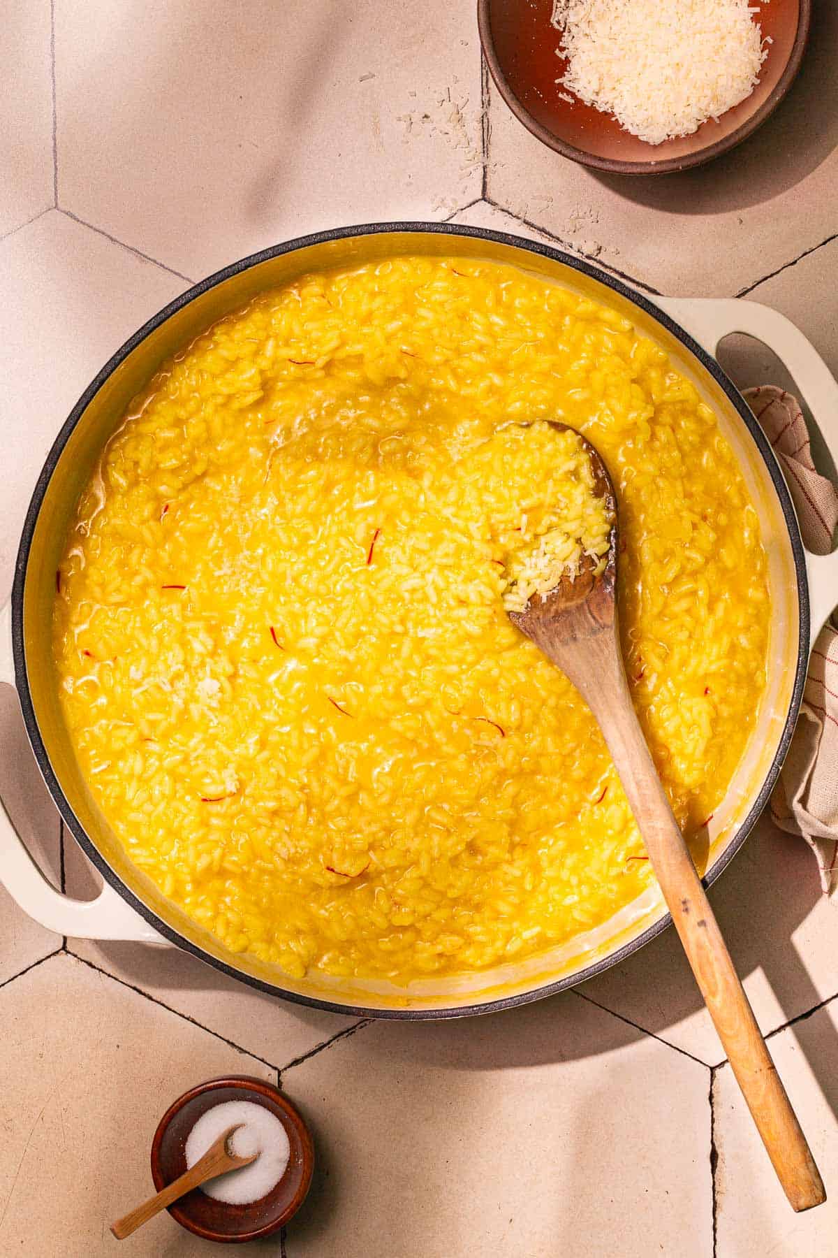 risotto alla milanese saffron risotto in a large pot being stirred with a wooden spoon next to a small bowl of salt with a spoon and a small bowl of grated parmesan cheese.