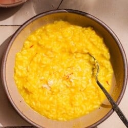 a bowl of risotto alla milanese saffron risotto with a spoon next to a small bowl of grated parmesan cheese and a glass of wine.