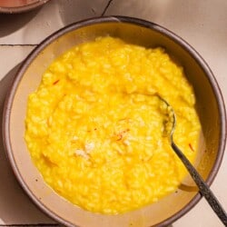 a bowl of risotto alla milanese saffron risotto with a spoon next to a small bowl of grated parmesan cheese and a glass of wine.