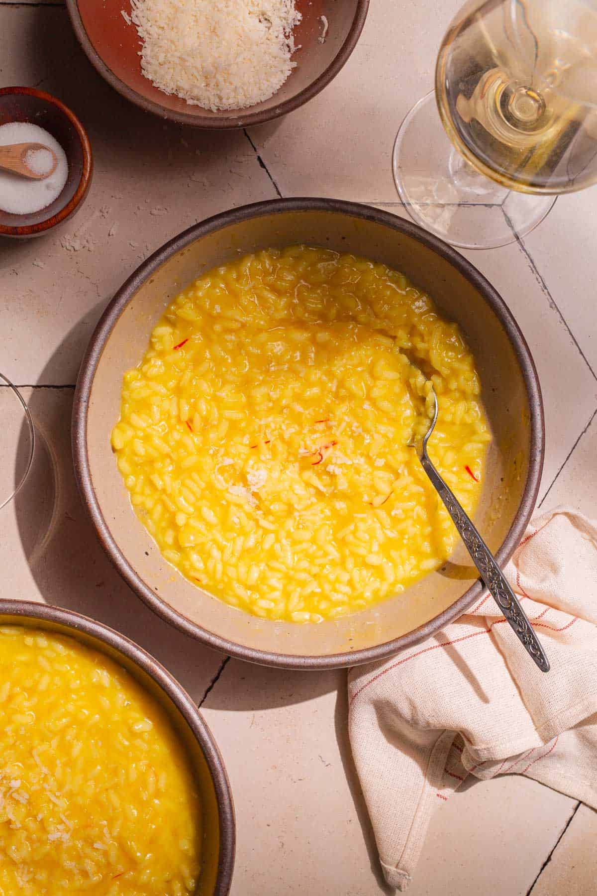 two bowls of risotto alla milanese saffron risotto next to a small bowl of grated parmesan cheese, a small bowl of salt, and a glass of wine.