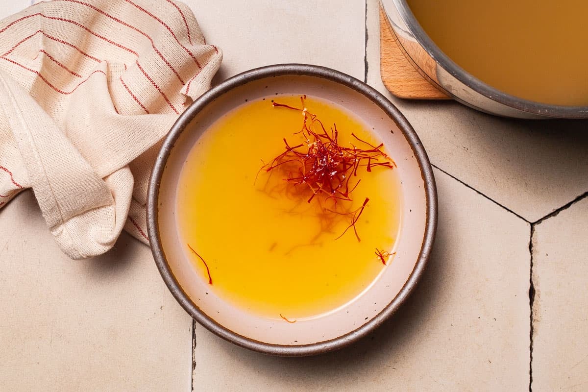 saffron threads being steeped in a bowl of hot broth, next to a pot of hot broth and a cloth napkin.