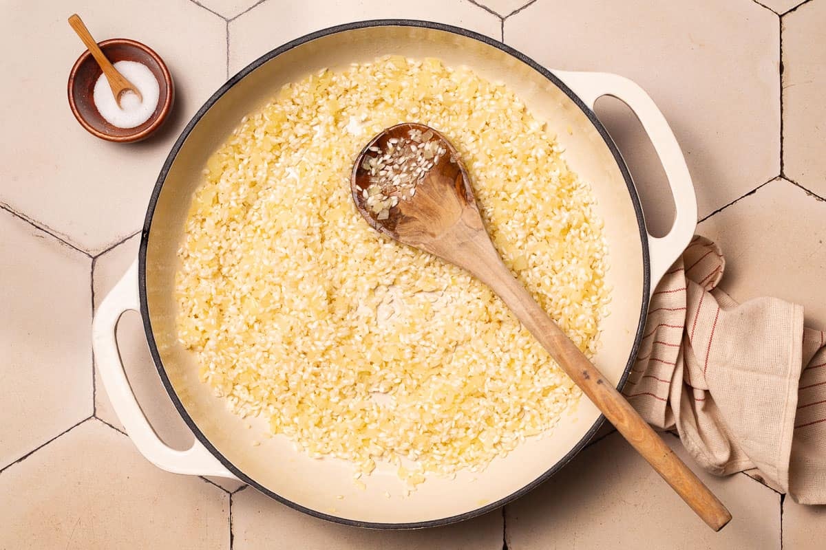 chopped onions and arborio rice being toasted in a pot with a wooden spoon next to a small bowl of salt with a spoon.