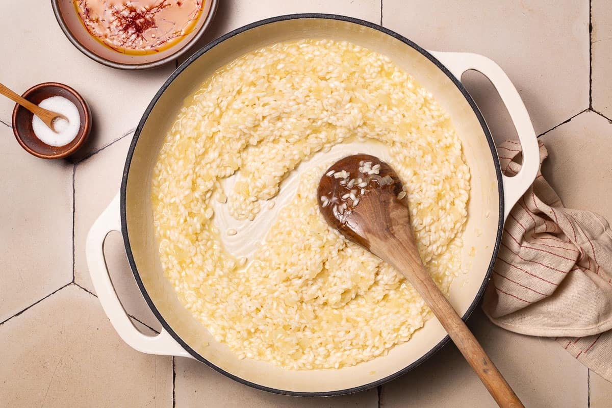 chopped onions and arborio rice being toasted in a pot with a wooden spoon next to a small bowl of salt with a spoon next to a bowl of saffron steeping in broth and a small bowl of salt with a spoon.