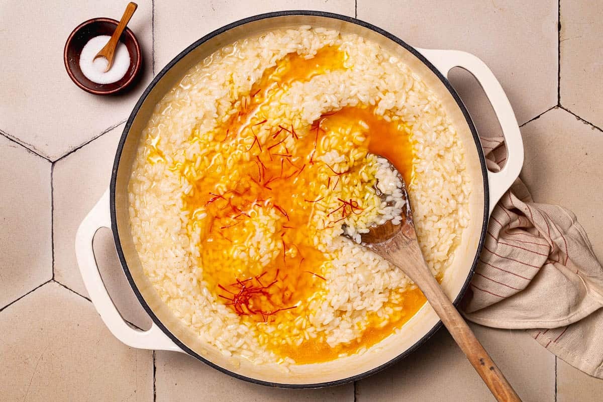 saffron and broth being added to the arborio rice in a large pot with a wooden spoon next to a small bowl of salt with a spoon.