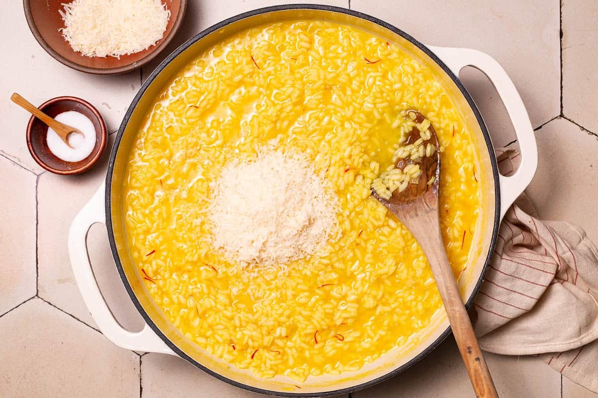 risotto alla milanese saffron risotto topped with parmesan cheese in a large pot being stirred with a wooden spoon next to a small bowl of salt with a spoon and a small bowl of grated parmesan cheese.