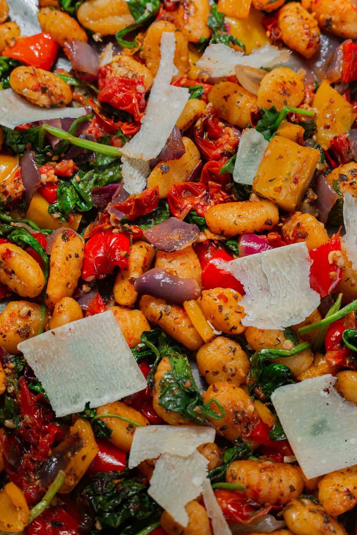 Close up of sheet pan gnocchi showing the bright red bell peppers, vibrant green spinach, assortment of spices, and large parmesan shavings on top.