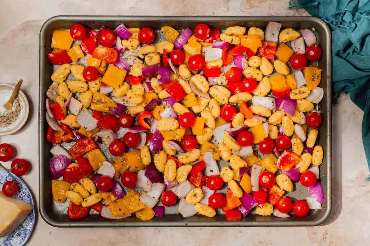 A sheet pan with gnocchi, red bell pepper, yellow bell pepper, red onion, cherry tomatoes, olive oil, oregano, basil, salt, thyme, red pepper flakes, and black pepper.