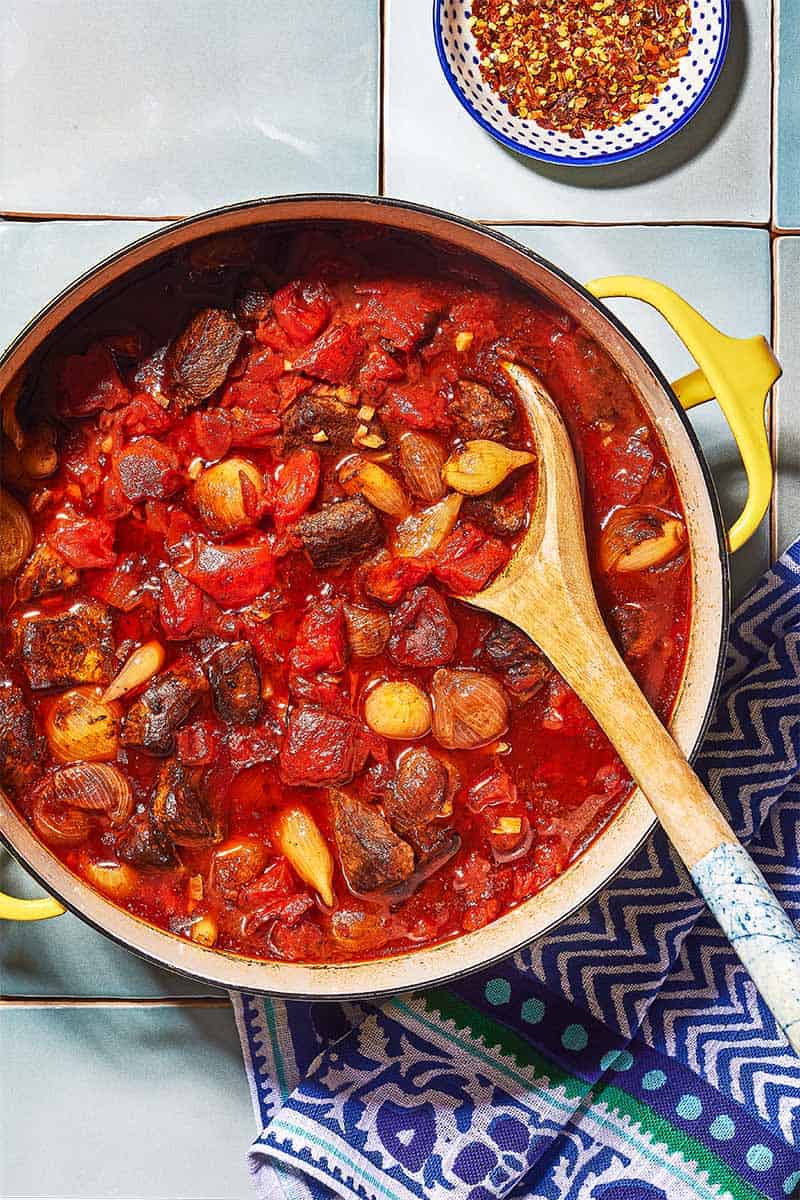 Overhead shot of greek beef stew with a wooden spoon, showing the tomato, onions, and beef.