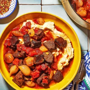 Overhead shot of greek beef stew in a bowl with mashed potatoes and red pepper flakes on the side.