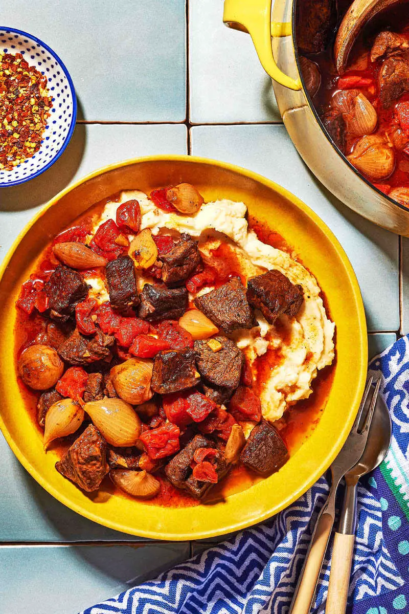 Overhead shot of greek beef stew in a bowl with mashed potatoes and red pepper flakes on the side.