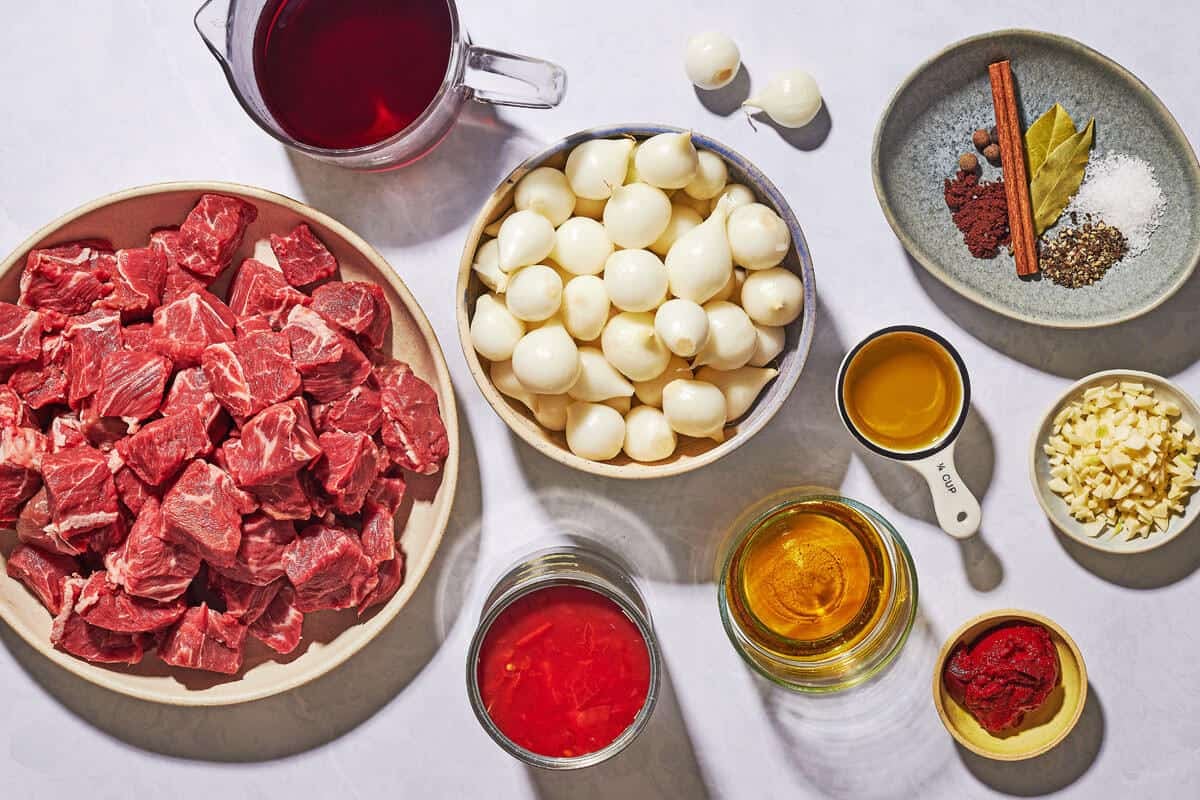 Ingredients for Greek beef stew, including chuck roast, salt, pepper, olive oil, peeled pearl onions, garlic cloves, tomato paste, bay leaves, cinnamon stick, clove, allspice berries, red wine, Cognac or Brandy, and canned tomatoes.