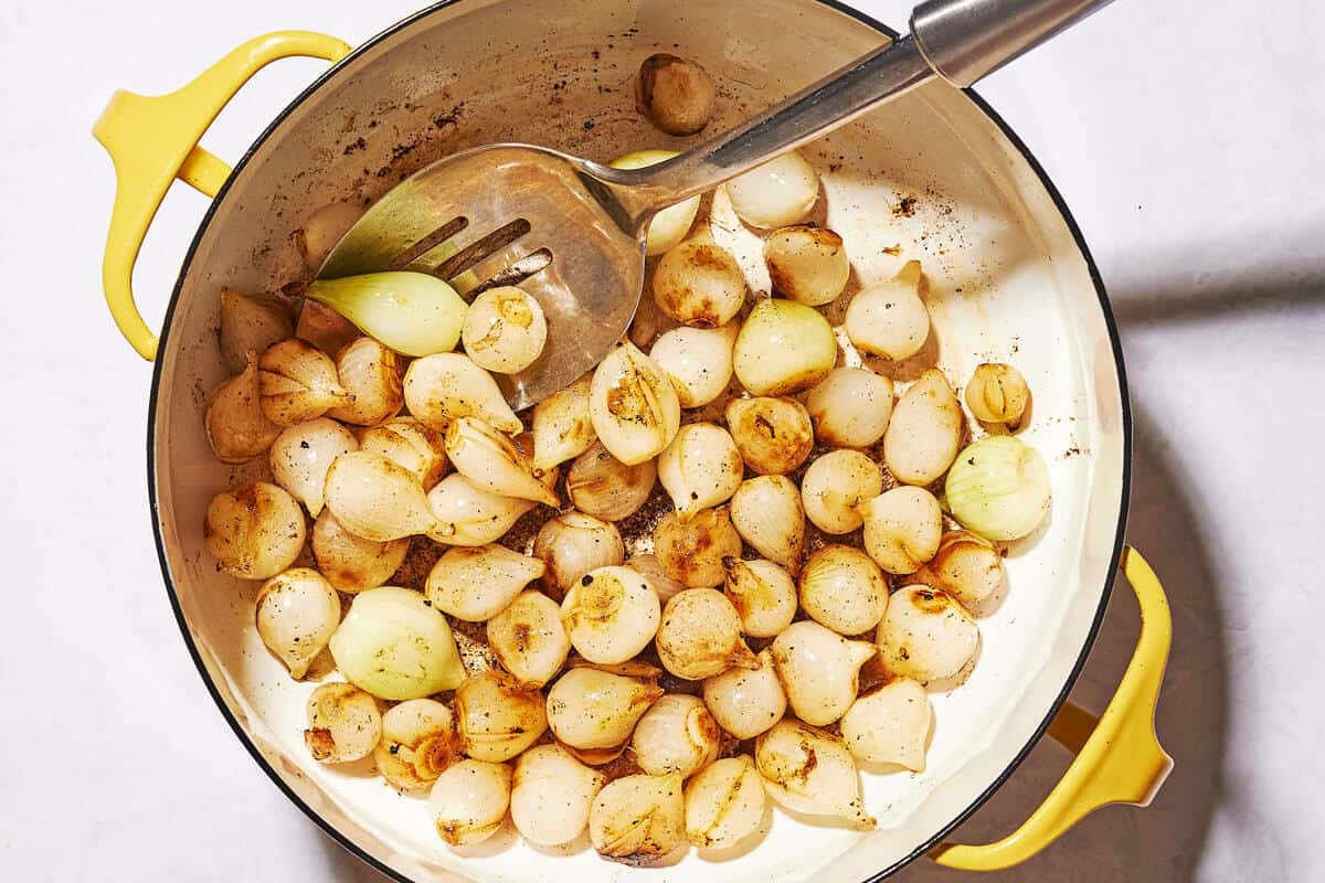 Pearl onions in a pot with a metal spoon, showing some browning in spots.