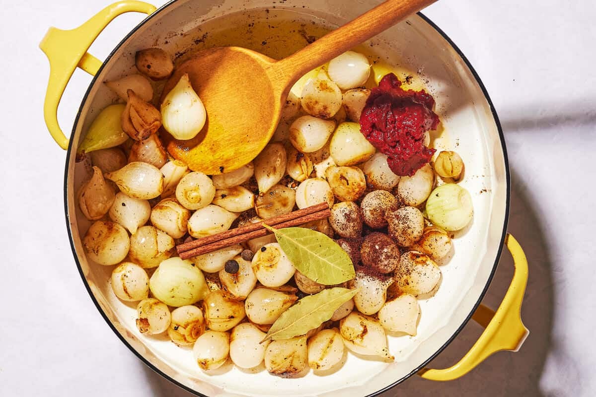 Peeled pearl onions in a pot with bay leaves and tomato paste that has not been stirred in yet.