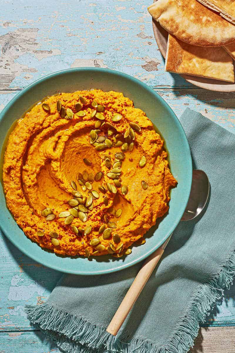 Overhead shot of pumpkin hummus with toasted pumpkin seeds and Aleppo pepper sprinkled over top and pita bread on the side.