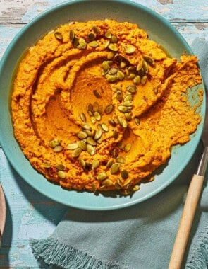 Overhead shot of pumpkin hummus with a spoon on the side. There is a smearing of hummus on the bowl, implying that someone has dipped pita into the hummus.
