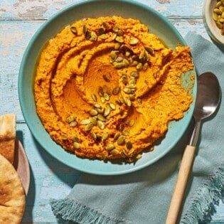 Overhead shot of pumpkin hummus with a spoon on the side. There is a smearing of hummus on the bowl, implying that someone has dipped pita into the hummus.