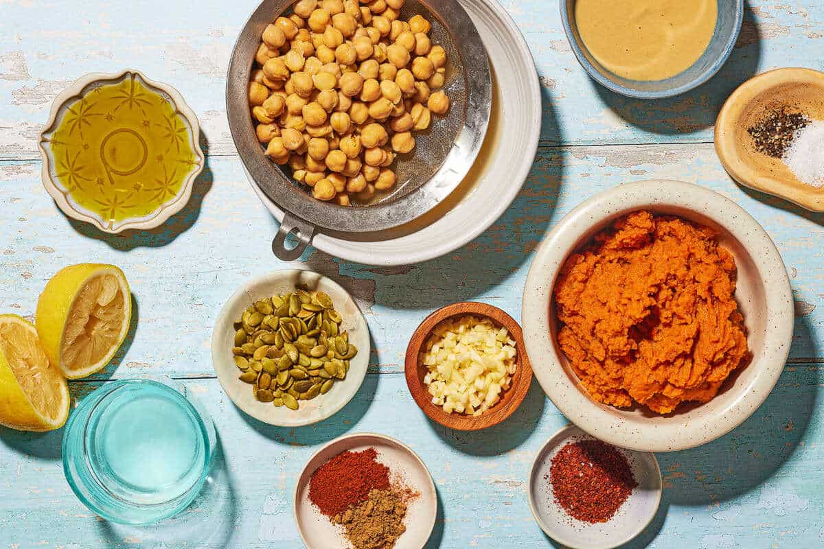 Ingredients for pumpkin hummus, including chickpeas, garlic, lemon, cumin, Aleppo pepper, olive oil, salt, pepper, tahini, pepitas, and pumpkin puree.