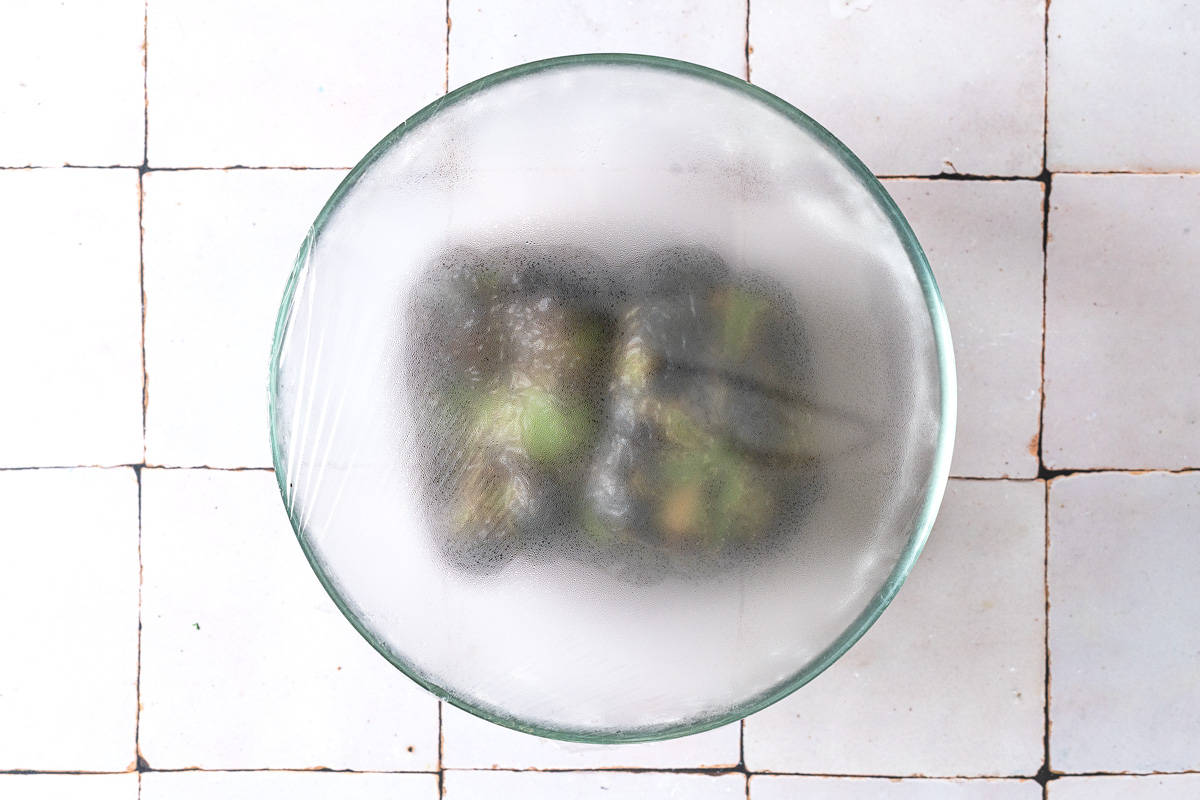 two charred green bell peppers in a bowl with plastic wrap on top, showing the steam being trapped in the bowl to soften the peppers.