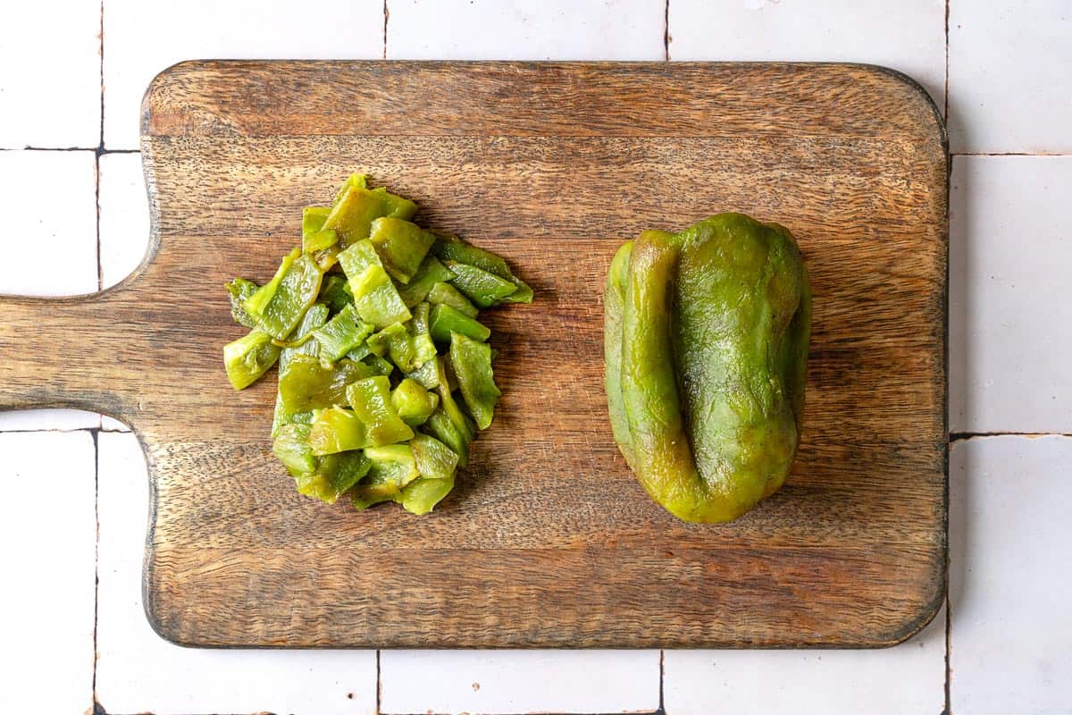 two roasted green bell peppers on a wooden cutting board. one is whole with the skin removed, showing the softened flesh, the other has been peeled and chopped into bite-sized pieces.
