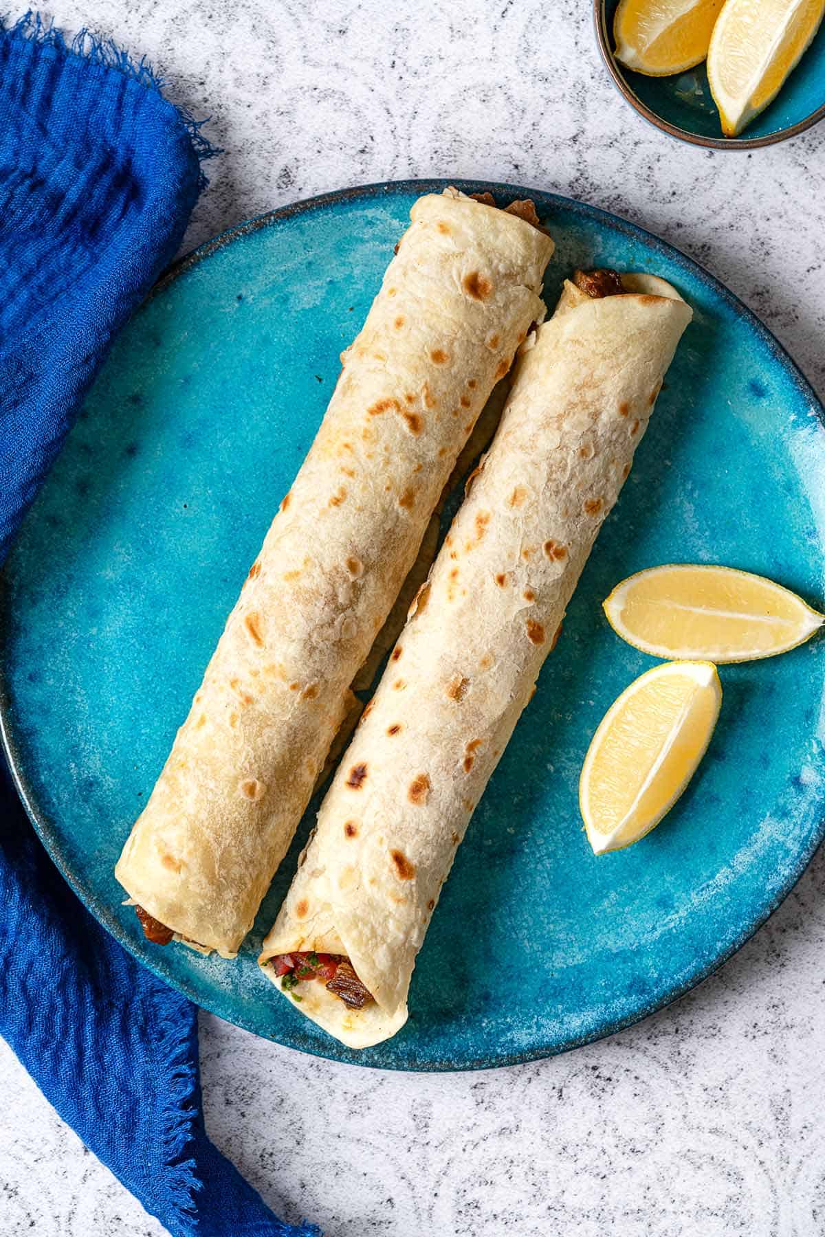 overhead shot of two tantuni steak wraps side by side on a plate with two lemon wedges on the side.