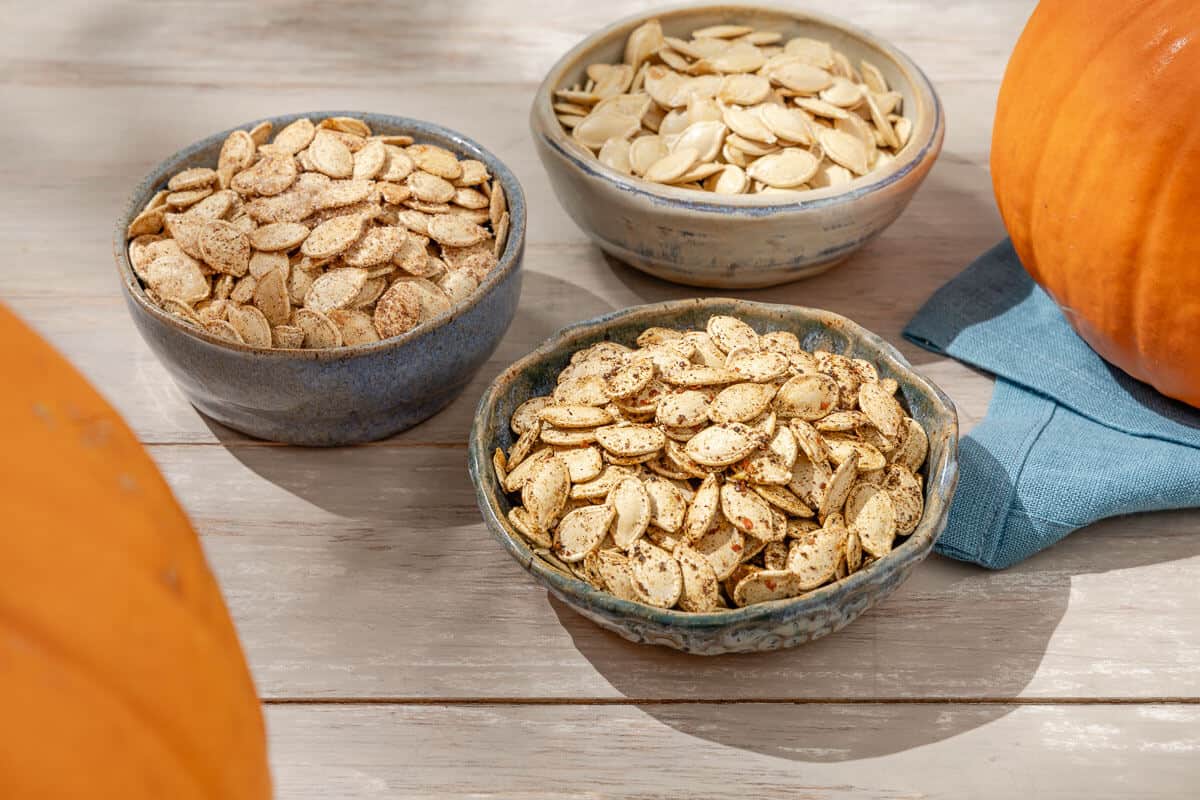 a photo of a bowl of sweet roast pumpkin seeds, a bowl of unseasoned pumpkin seeds and a bowl of savory pumpkin seeds in between two pumpkins.