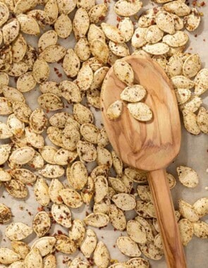 a close up of savory seasoned roast pumpkin seeds with a wooden serving spoon on a parchment lined sheet pan.