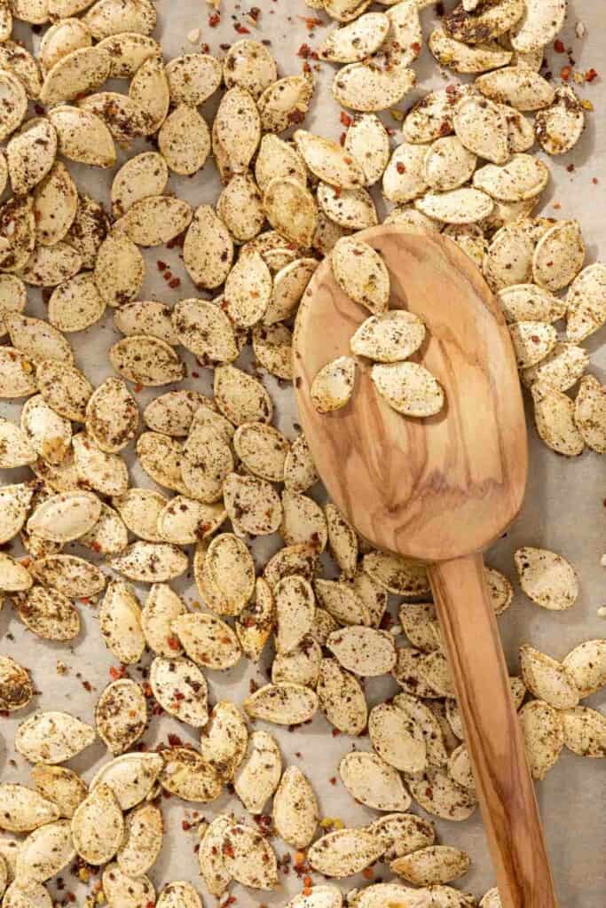 a close up of savory seasoned roast pumpkin seeds with a wooden serving spoon on a parchment lined sheet pan.