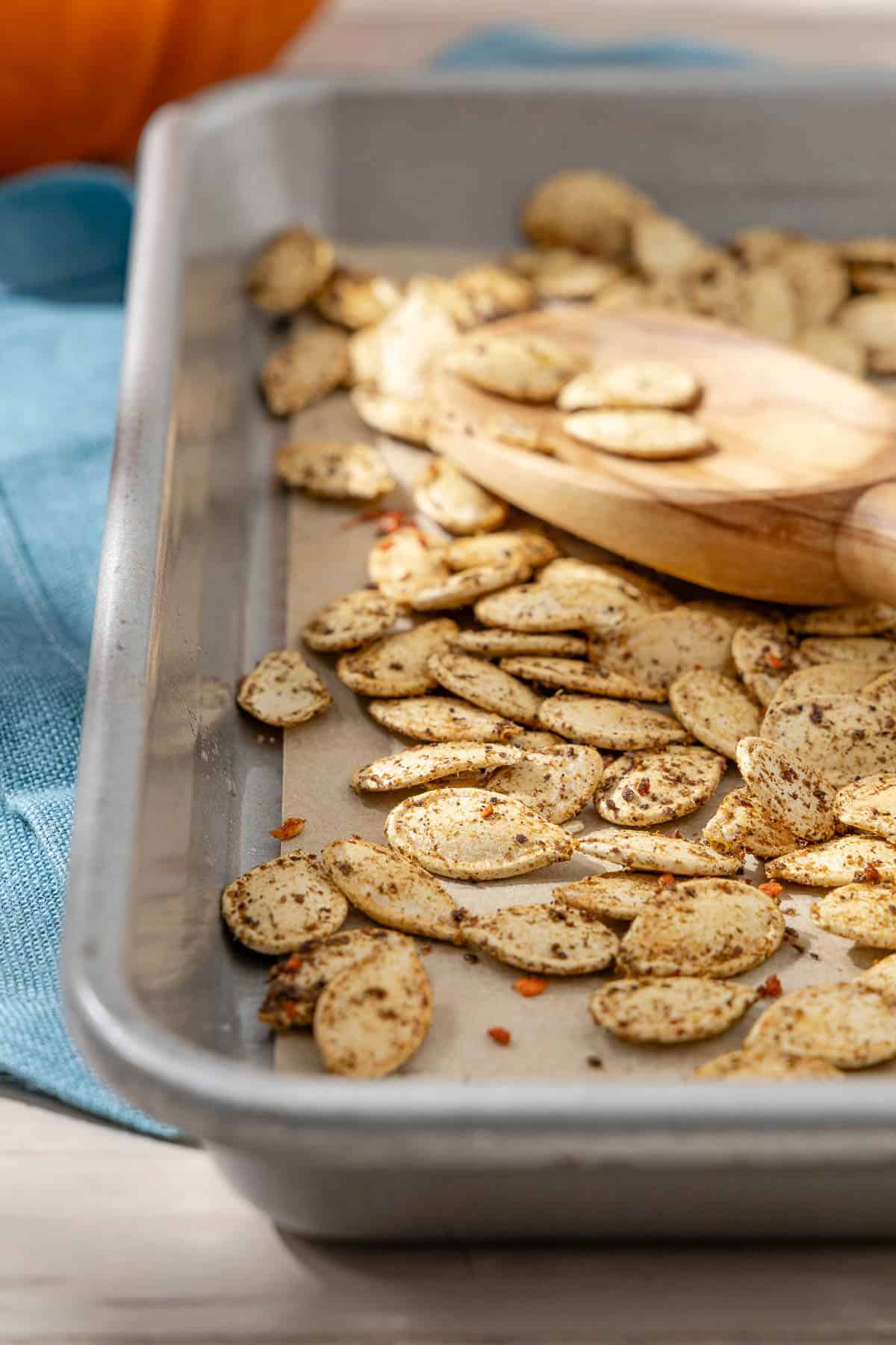 a roasted pumpkin seed recipe where there is a close-up photo of savory seasoned roasted pumpkin seeds with a wooden serving spoon on a parchment-lined sheet pan.