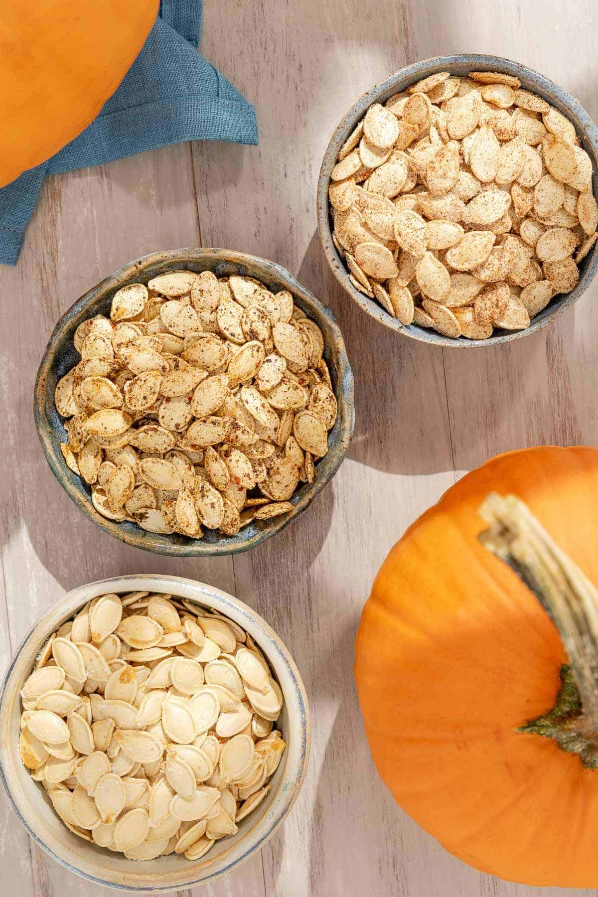 an overhead photo of a bowl of sweet roast pumpkin seeds, a bowl of unseasoned pumpkin seeds and a bowl of savory pumpkin seeds in between two pumpkins.