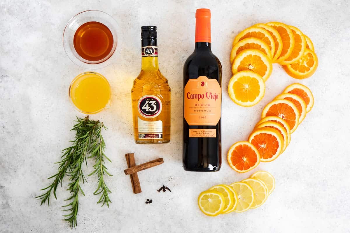 ingredients for holiday sangria including oranges, lemon, cinnamon sticks, rosemary sprigs, Licor 43, honey, black peppercorns, cloves, and a bottle of red wine.