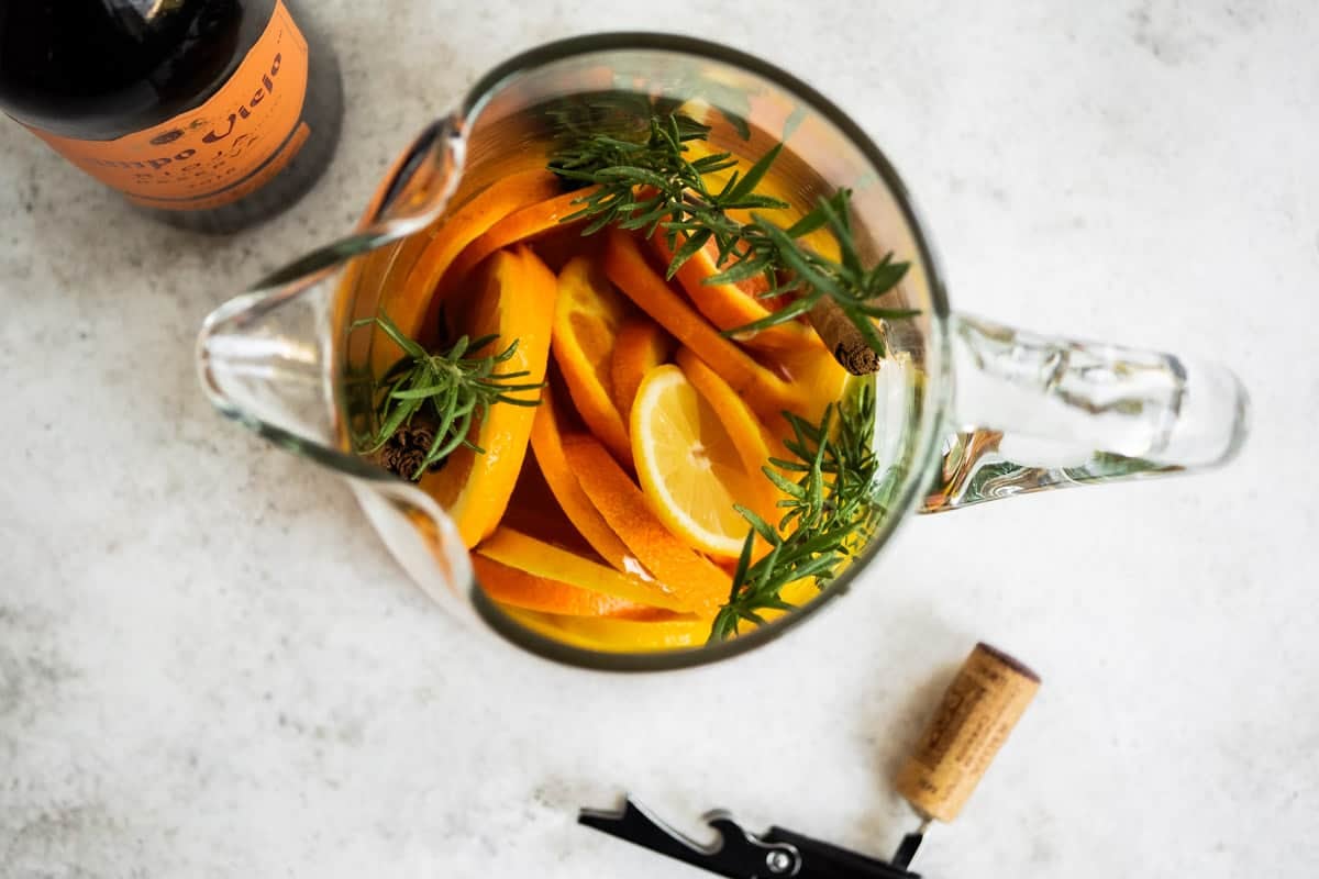 an overhead photo of holiday sangria garnished with orange wheels and sprigs of rosemary next to a bottle of red wine, and a corkscrew with a cork.