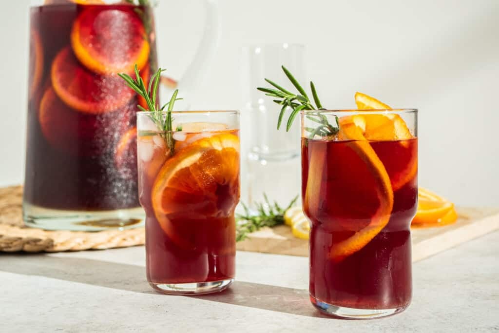 a close up of two glasses of holiday sangria garnished with orange wheels and sprigs of rosemary in front of a pitcher of holiday sangria.