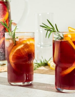 a close up of two glasses of holiday sangria garnished with orange wheels and sprigs of rosemary in front of a pitcher of holiday sangria.