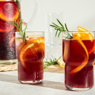 a close up of two glasses of holiday sangria garnished with orange wheels and sprigs of rosemary in front of a pitcher of holiday sangria.