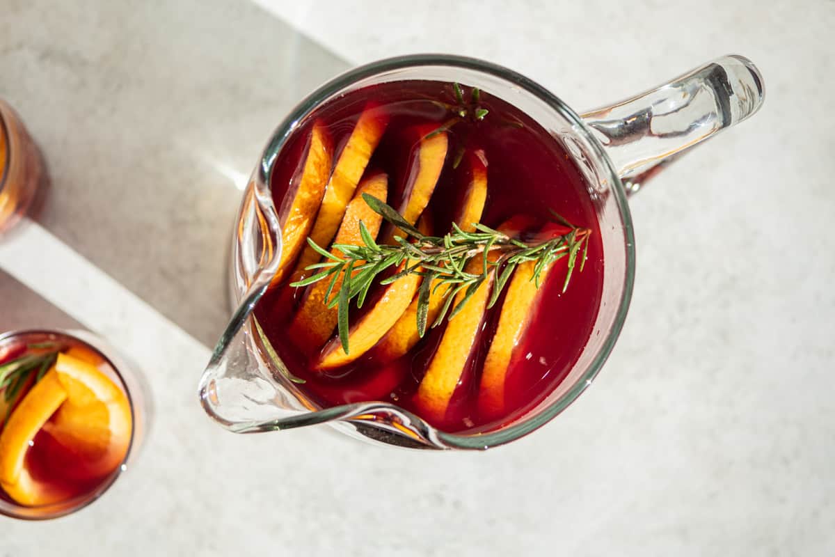 an overhead photo of holiday sangria garnished with orange wheels and sprigs of rosemary next to a glass of holiday sangria.