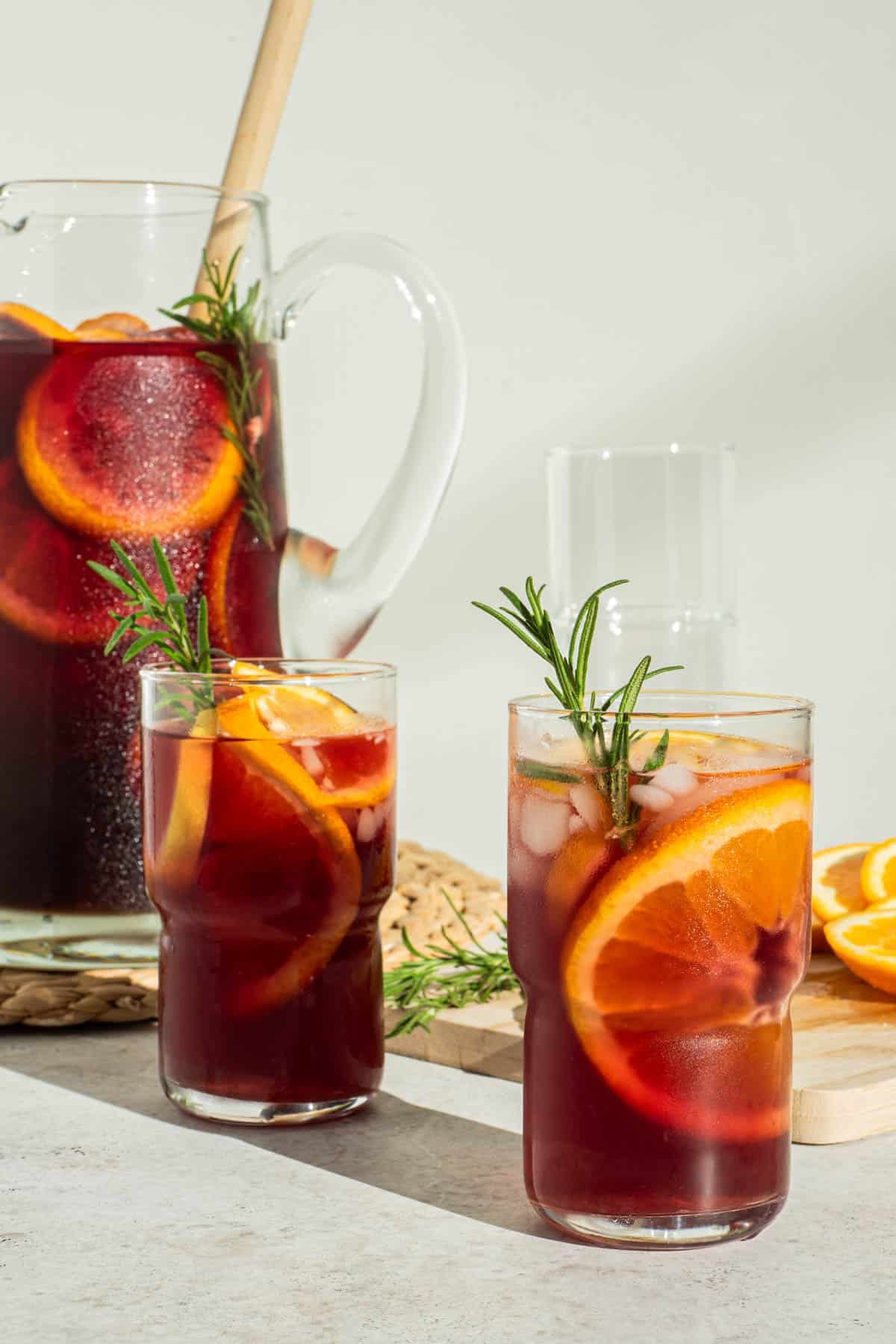 two glasses of holiday sangria garnished with orange wheels and sprigs of rosemary in front of a pitcher of holiday sangria with a wooden stirring spoon.