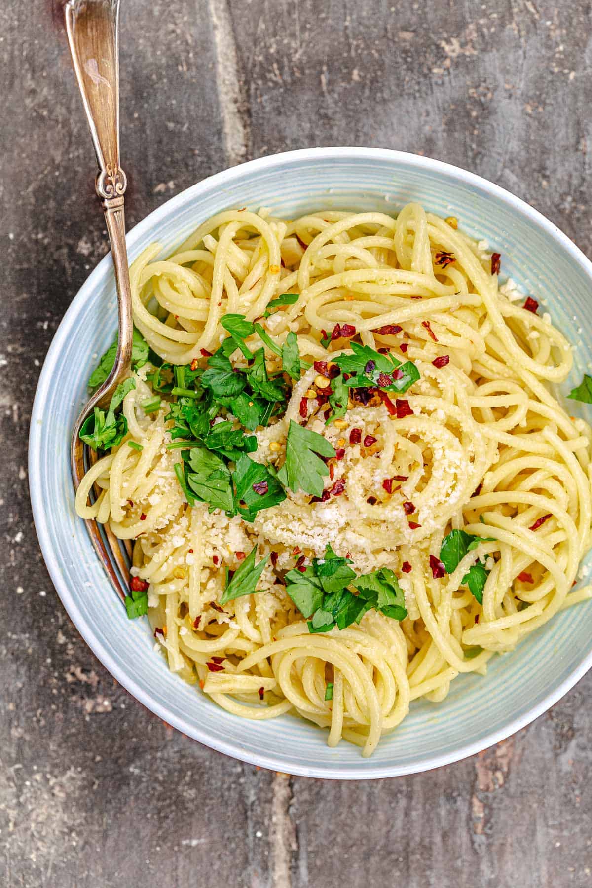 an overhead photo of spaghetti aglio e olio topped with grated parmesan, red pepper flakes and parsley in a bowl with a fork.